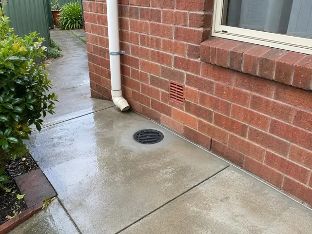 Outdoor concrete path beside brick wall with a round drain grate and downpipe in Rydalmere